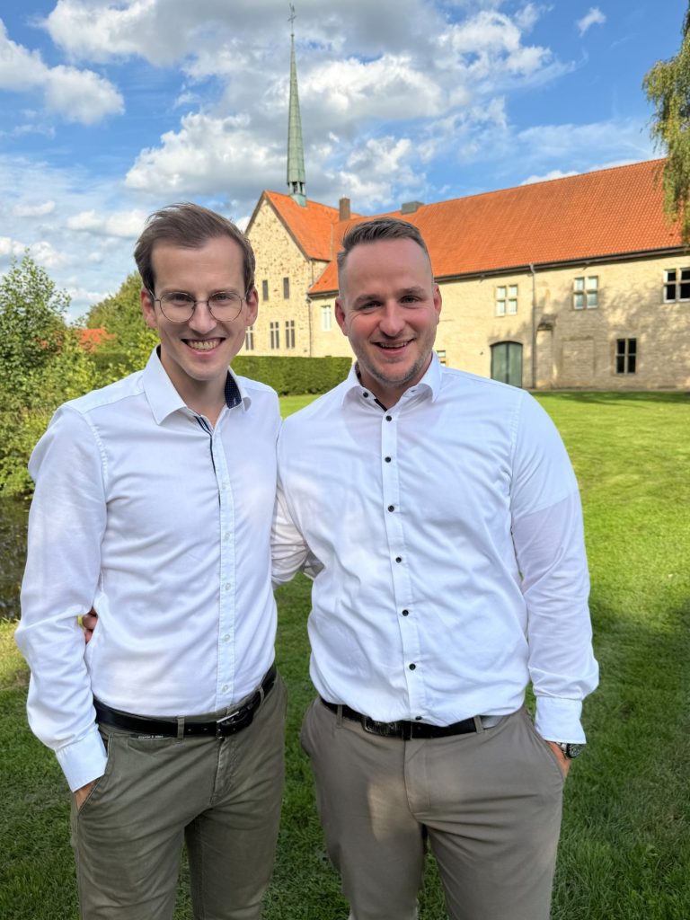 Fabian & Heiner Zwei lächelnde Männer stehen vor einem roten Fahrzeug.